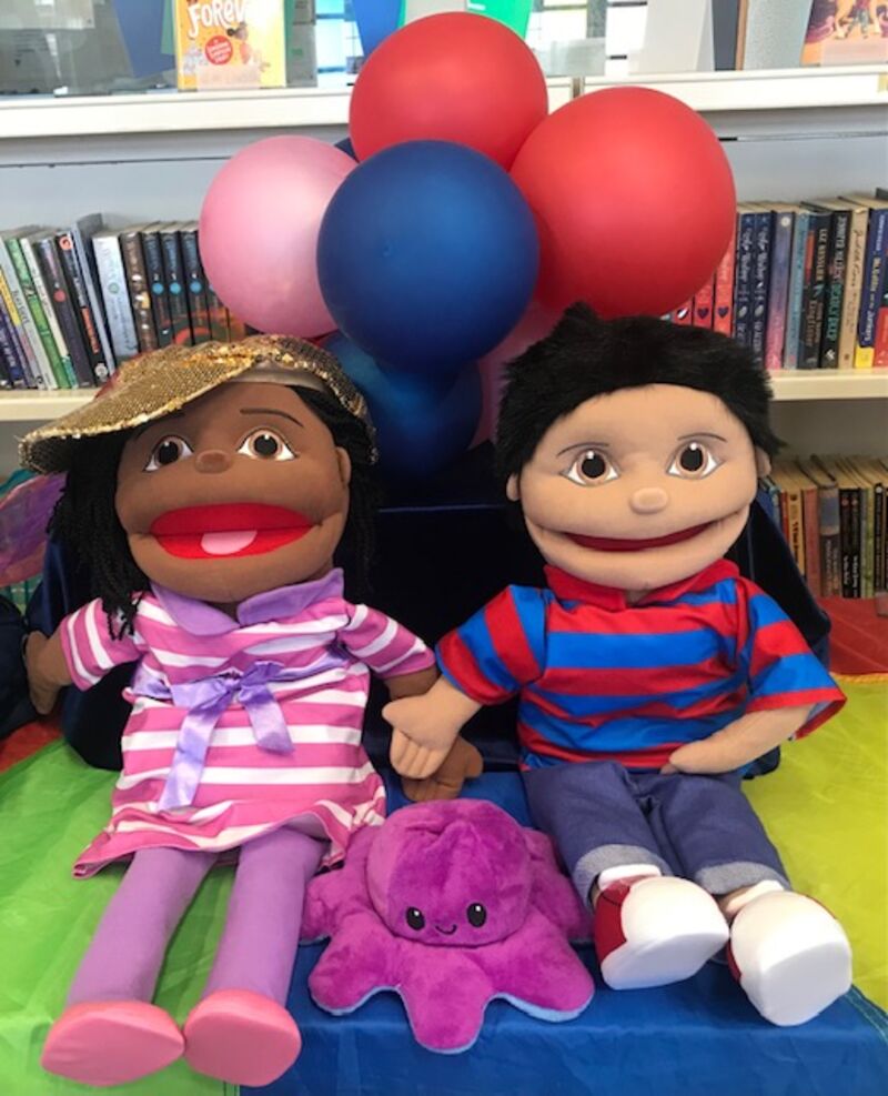Girl puppet in a white and purple striped dress sitting next to boy puppet dresses in a red and blue striped shirt and blue jeans. With balloons behind them.