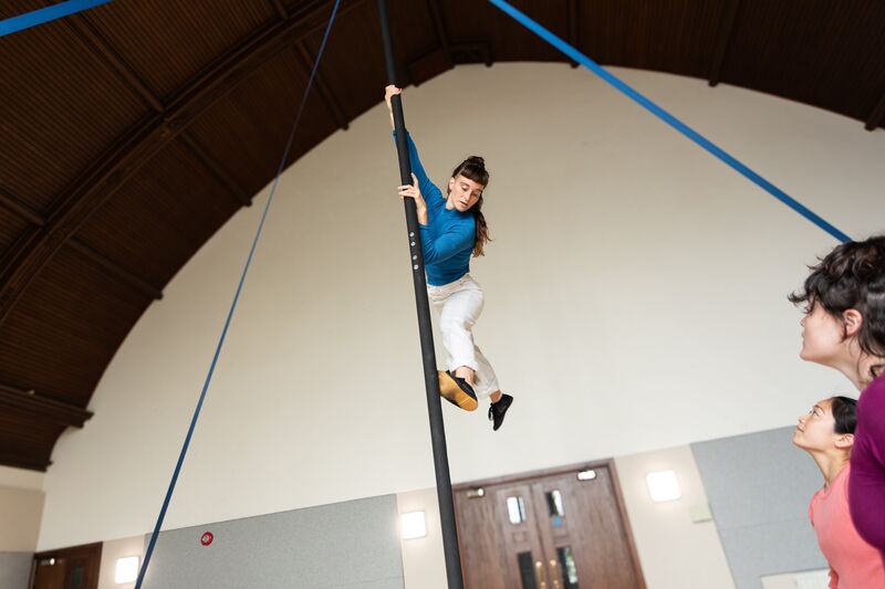 A woman climbs a vertical pole, and has one leg out behind her