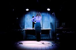 Liza Bec, tall, white, with long brown hair, stands with their left hand on their hip facing three large blackboards. A piece of chalk is in their hand, poised to write. They wear a fitted purple top and wide black trousers. Two spotlights illuminate the scene.
