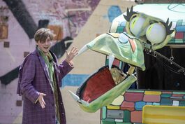 A big green dragon puppet head is singing. A man in a purple coat holds out his hands to introduce the dragon..