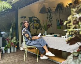 A Brunette Woman With Glasses Sits Smiling At A Table Surrounded By Flowers