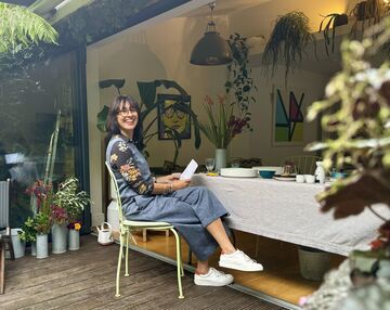 A Brunette Woman With Glasses Sits Smiling At A Table Surrounded By Flowers