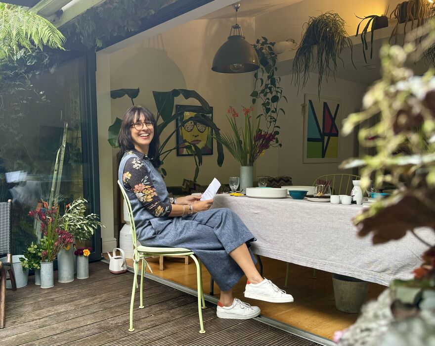 A Brunette Woman With Glasses Sits Smiling At A Table Surrounded By Flowers