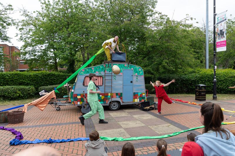 A cute brightly painted retro caravan.  Three performers in pyjamas are throwing a golden egg to each other.  One actor is on top of the caravan.