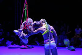     Two performers have their back to us, illuminated in purple light. One person is suspended in air by rope bondage knot, with neon red, pink and green rope, his hands crossed behind his back. The other has his back to the camera and is gently holding the first person's head and pressing their own to it, creating a sense of connection and safety. In the purple background various mundane objects are spun in neon macramé creating unique patterns based on their shapes. An audience is sitting on the floor, watching. 