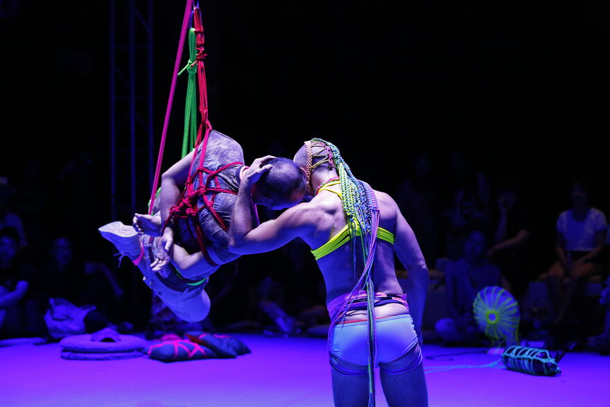     Two performers have their back to us, illuminated in purple light. One person is suspended in air by rope bondage knot, with neon red, pink and green rope, his hands crossed behind his back. The other has his back to the camera and is gently holding the first person's head and pressing their own to it, creating a sense of connection and safety. In the purple background various mundane objects are spun in neon macramé creating unique patterns based on their shapes. An audience is sitting on the floor, watching. 