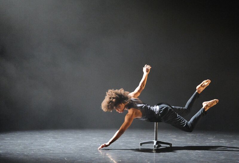 A dancer lays across a stool and reaches one hand down towards the floor.