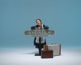 A young dancers dressed as Anne of Green Gables with ginger hair and two plaits leading on train station sign for Avonlea, with suitcases next to her. 