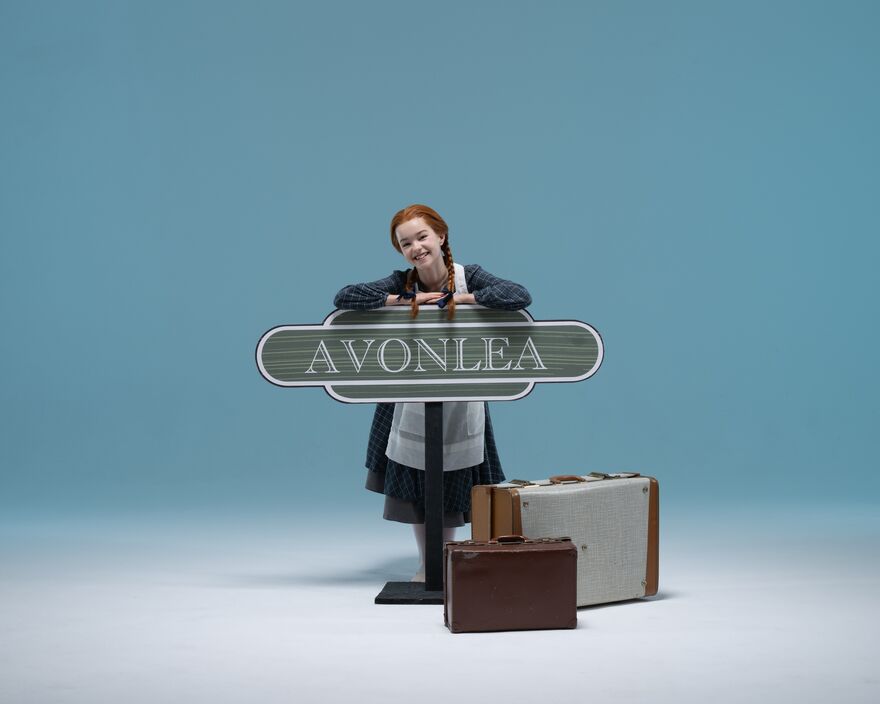 A young dancers dressed as Anne of Green Gables with ginger hair and two plaits leading on train station sign for Avonlea, with suitcases next to her. 