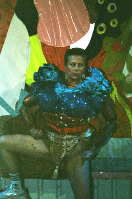 An artist dressed in brightly coloured carnival style clothing posing with their leg up