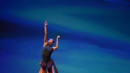 Female dancer is lifted in the air while reaching up with one arm. There is a blue background behind her.
