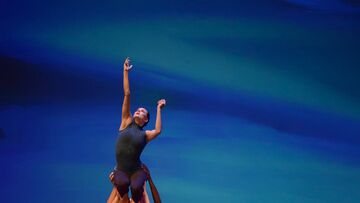 Female dancer is lifted in the air while reaching up with one arm. There is a blue background behind her.