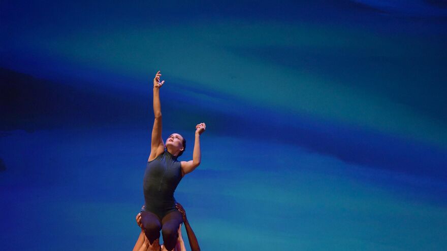 Female dancer is lifted in the air while reaching up with one arm. There is a blue background behind her.