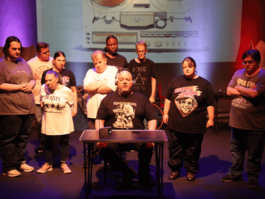 Everyone in the group is dressed in t-shirts featuring iconic music artists or bands, including The Beatles, Elton John, Queen, Guns N’ Roses, Iron Maiden, and Pink Floyd. Behind them, a large image of a vintage TEAC reel-to-reel tape recorder is projected onto a screen. The stage lighting is moody with purple and red hues, adding to the dramatic tone of the scene.