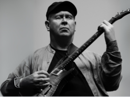 This black-and-white photo depicts Adrian Lee, a white man playing an electric guitar. Adrian’s figure fills the frame from just below the chest up. Adrian faces the camera squarely but his eyes are shut with deep concentration as he plays his guitar which is held high to his chest across his left shoulder. His right hand fingers pluck the string while his left hand fingers are curled to press the strings on the guitar’s neck. He wears a dark baseball cap, a light-coloured shirt with a mandarin colour unbuttoned over a dark t-shirt. The background is light, and Adrian’s stance is dramatic and strong and the image of him is clearly articulated in this black-and-white photo.