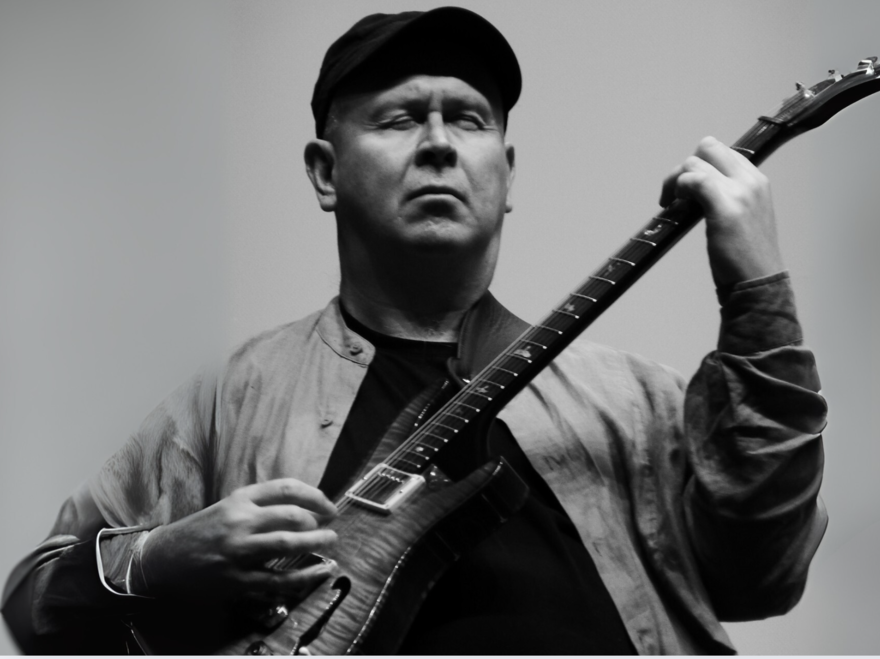 This black-and-white photo depicts Adrian Lee, a white man playing an electric guitar. Adrian’s figure fills the frame from just below the chest up. Adrian faces the camera squarely but his eyes are shut with deep concentration as he plays his guitar which is held high to his chest across his left shoulder. His right hand fingers pluck the string while his left hand fingers are curled to press the strings on the guitar’s neck. He wears a dark baseball cap, a light-coloured shirt with a mandarin colour unbuttoned over a dark t-shirt. The background is light, and Adrian’s stance is dramatic and strong and the image of him is clearly articulated in this black-and-white photo.