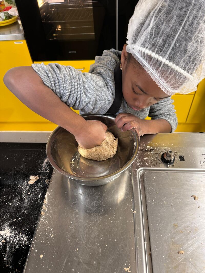 A young girl wearing a catering hair net and apron is standing holding and looking at a stainless steel bowl which contains dough. She needs the dough with her fist.