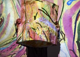 The image shows artist Chandrakant Patel holding up his painting artwork within a projection