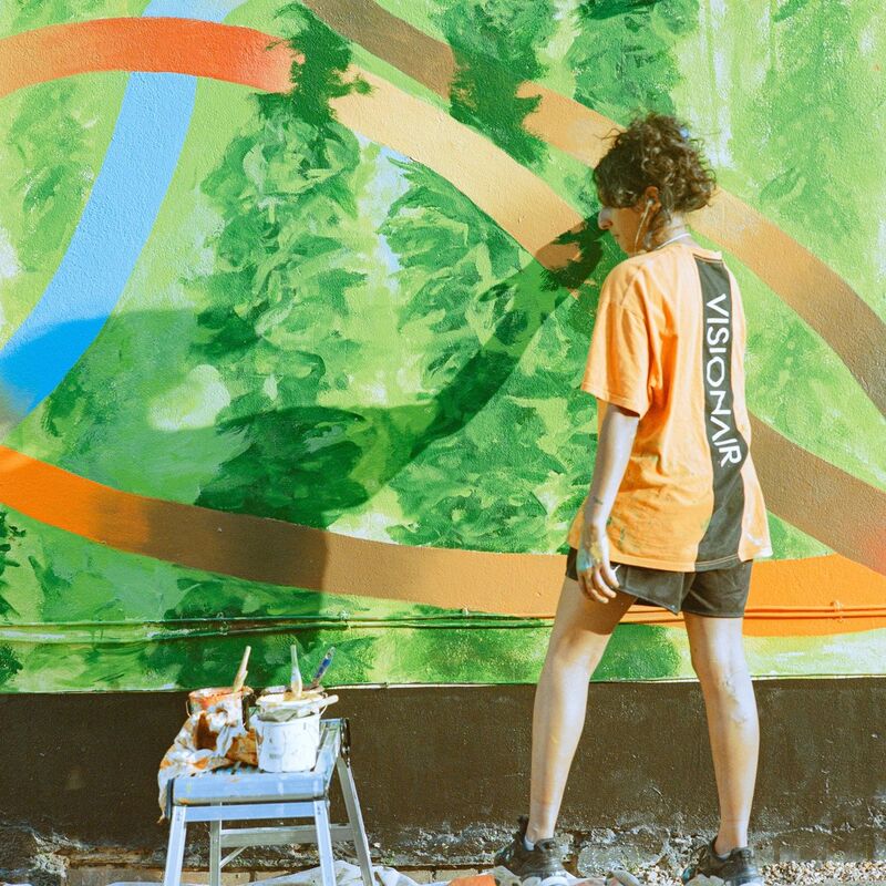 Shiza standing and painting the Hop Pole Mural, with some pots of paints on a stool next to her