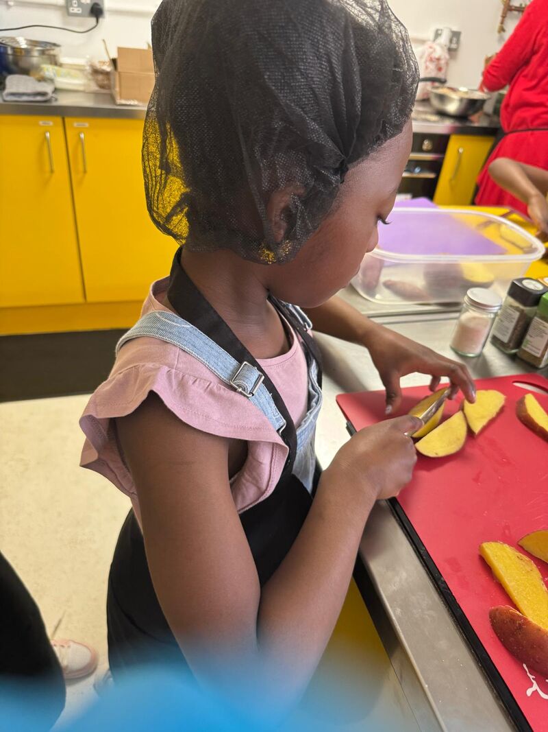 A young girl wearing a catering hair net and apron is standing holding a knife and carefully  slices mango. She is in a kitchen with iother childen and is using a red chopping board, where other slices are randomly placed