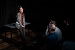  woman with long brown hair is wearing a brown turtle neck and a blazer, leaning against a black table and smiling dreamily into the distance. She is stood in front of an audience, we can see two people sat down watching her. 