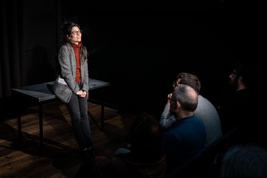  woman with long brown hair is wearing a brown turtle neck and a blazer, leaning against a black table and smiling dreamily into the distance. She is stood in front of an audience, we can see two people sat down watching her. 