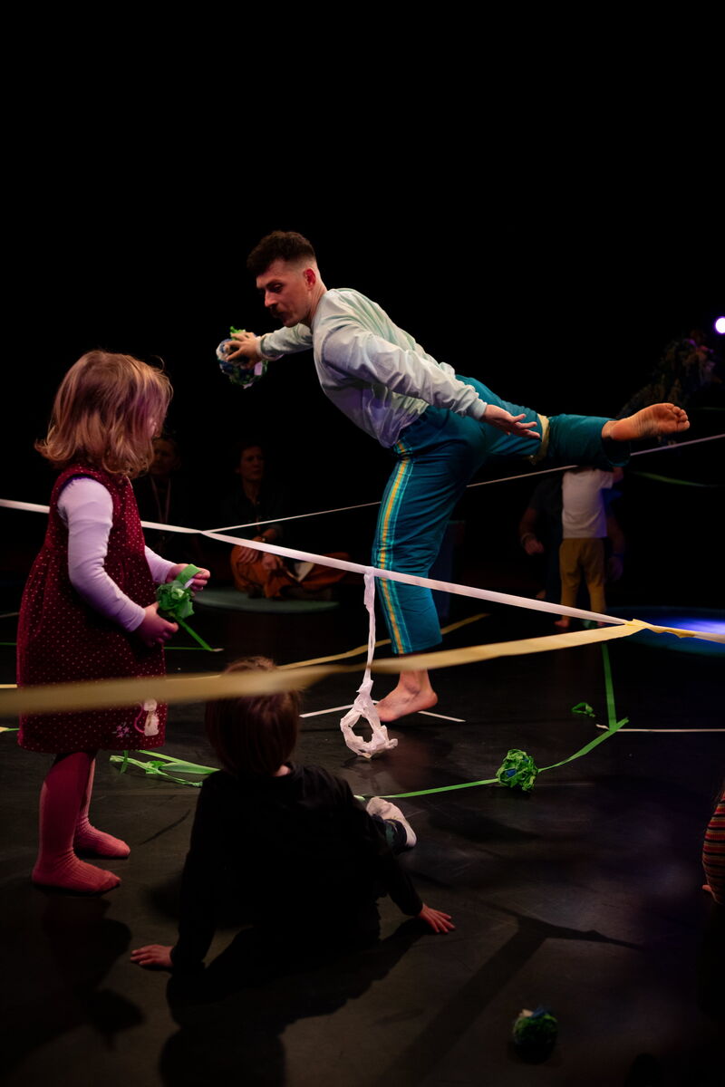 A male dancer stands on one leg while a young girl watches. There is a line of sticky tape stretched between them. 