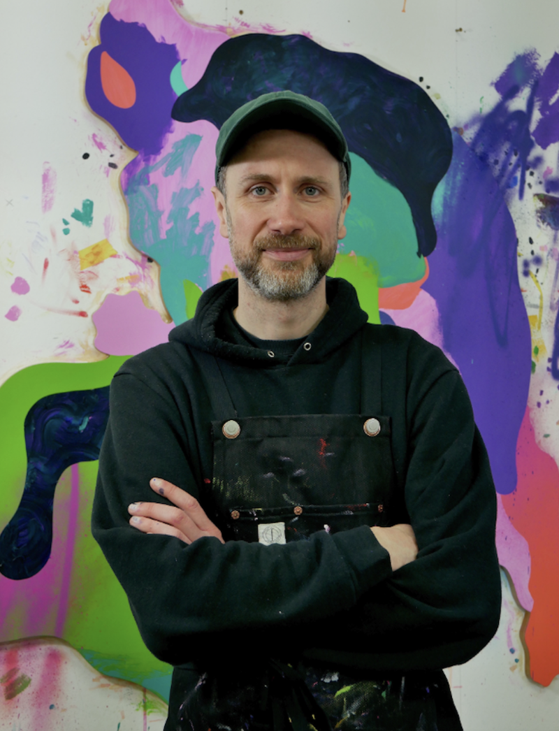 A man folds his arms and smiles at the camera in front of a painting piece