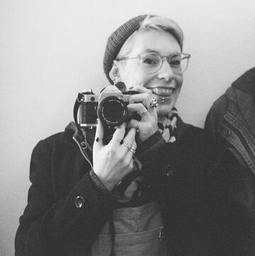 Black And White Image Of A Photographer Wearing A Hat, Black Coat, And Clear Framed Glasses Who Is Smiling And Holding A Camera To The Lower Left Of Their Face