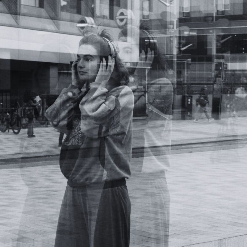 A black and white photograph of a person wearing headphones, standing in an urban setting with glass reflections creating a layered, ghostly effect. The background features a city street with pedestrians, bicycles, and a modern building with large windows and a London Underground sign.