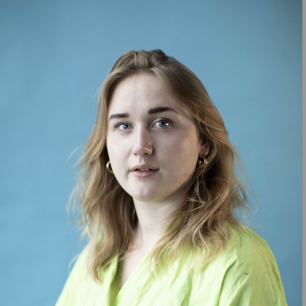 Kate Andrews, a white femme creative in her late 20s, looks directly into the camera. She is wearing a loose brat green collared shirt and gold earrings, and her dark blonde wavy hair is down around her shoulders.