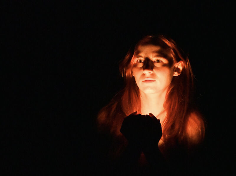 A woman stands in darkness. Her face is lit by a warm light that she holds in her hands.