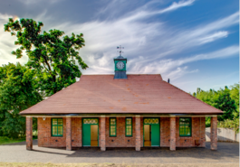 Photograph of a red brick building with a green clock tower on the roof. There are green doors and windows, and 6 pillars in front. The sky is blue with white clouds.  