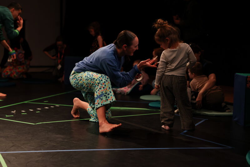 A female dancer kneels in front of a young girl holding out sticky tape towards her in an invitation for her to tape