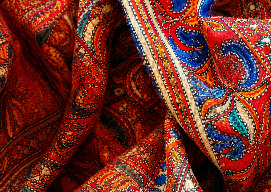 Close-up of a richly patterned red saree fabric with folds, showing detailed blue, teal, orange and cream motifs inspired by traditional textile design.