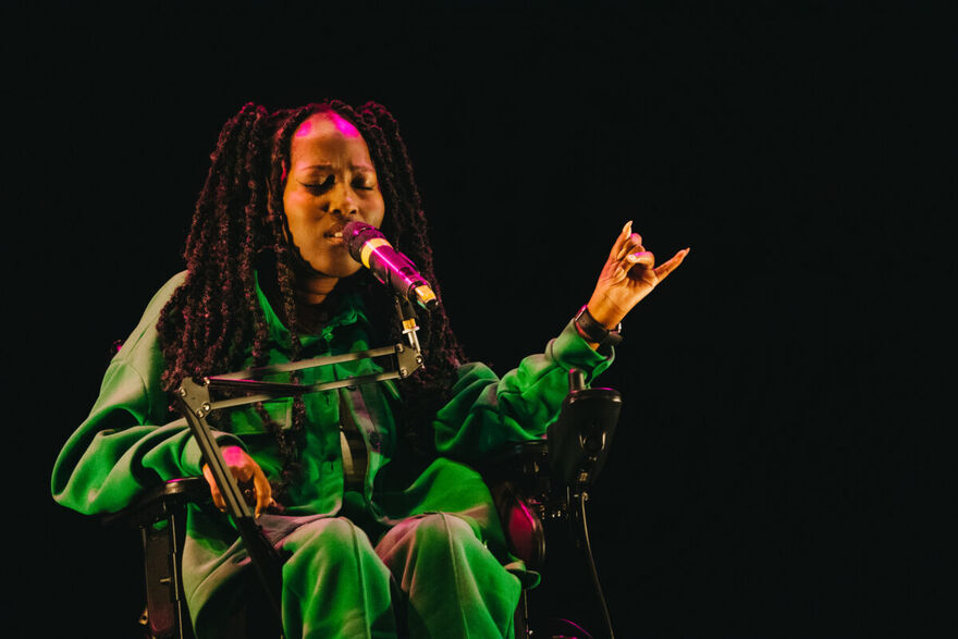 Miss Jacqui performs onstage. She sings into a microphone, hand half raised with her eyes shut.