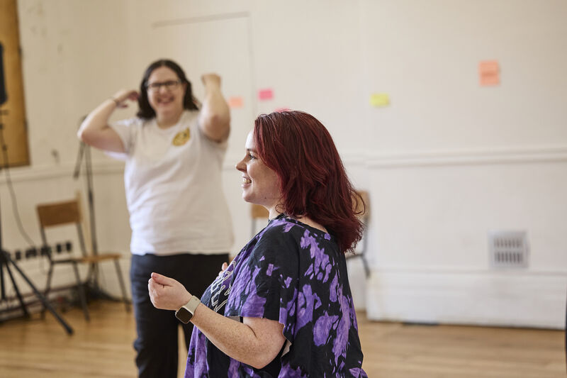 Ruth smiles in the forground in purple with red hair, a participant in a white t-shirt smiles in the background. Post its cover the wall behind them in a workshop