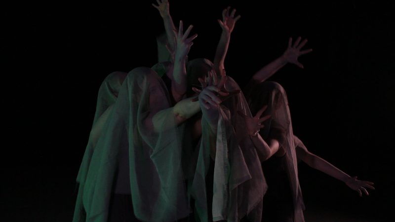 a group of dancers stand close together and reach their arms out in different directions. Their faces are covered by gray veils.