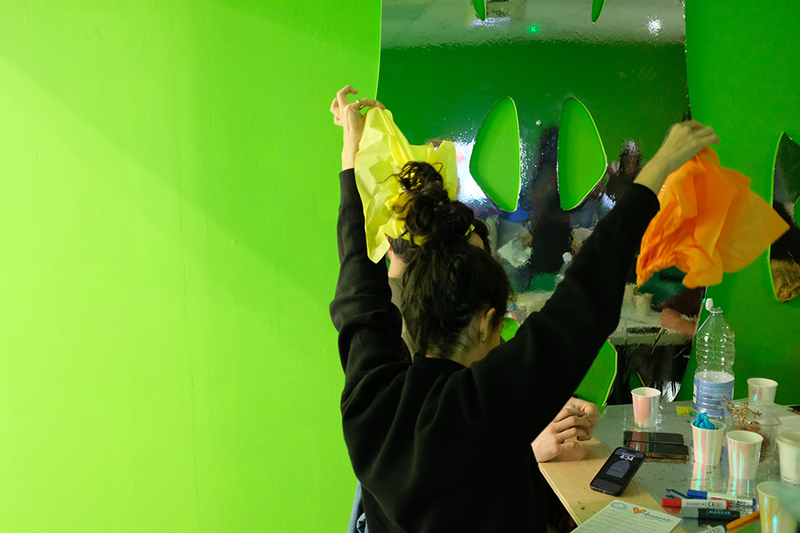 A participant creating a hat for the The Elysium Gaming Workshop, holding up a piece of orange and a piece of yellow paper above their head