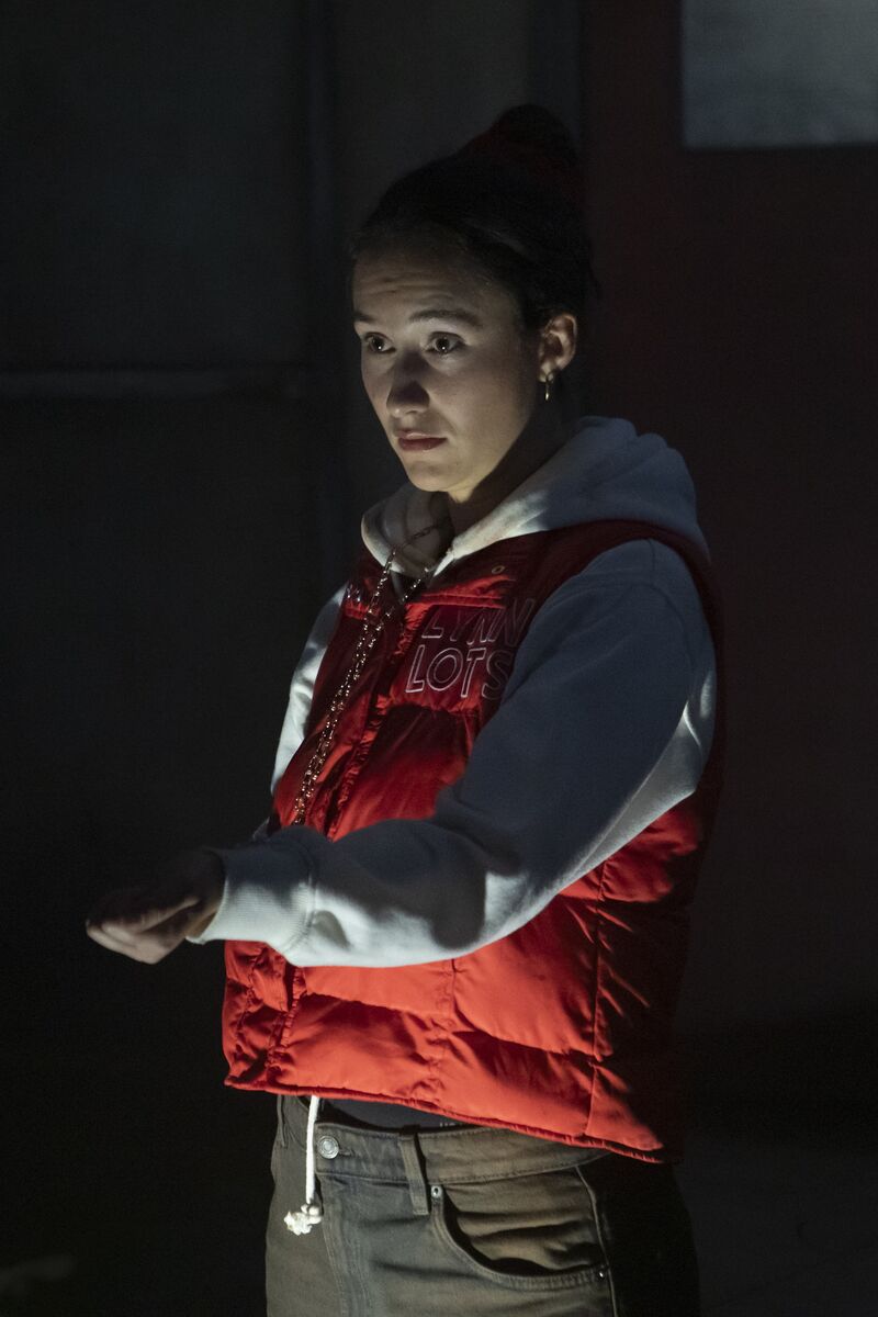 A woman in an orange and white jacket holds out her left hand. She looks scared. The lighting is dark and up-lit.