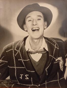 Photographic portrait of Jack Stanford smiling into camera with a hat, bow tie, and check coat on