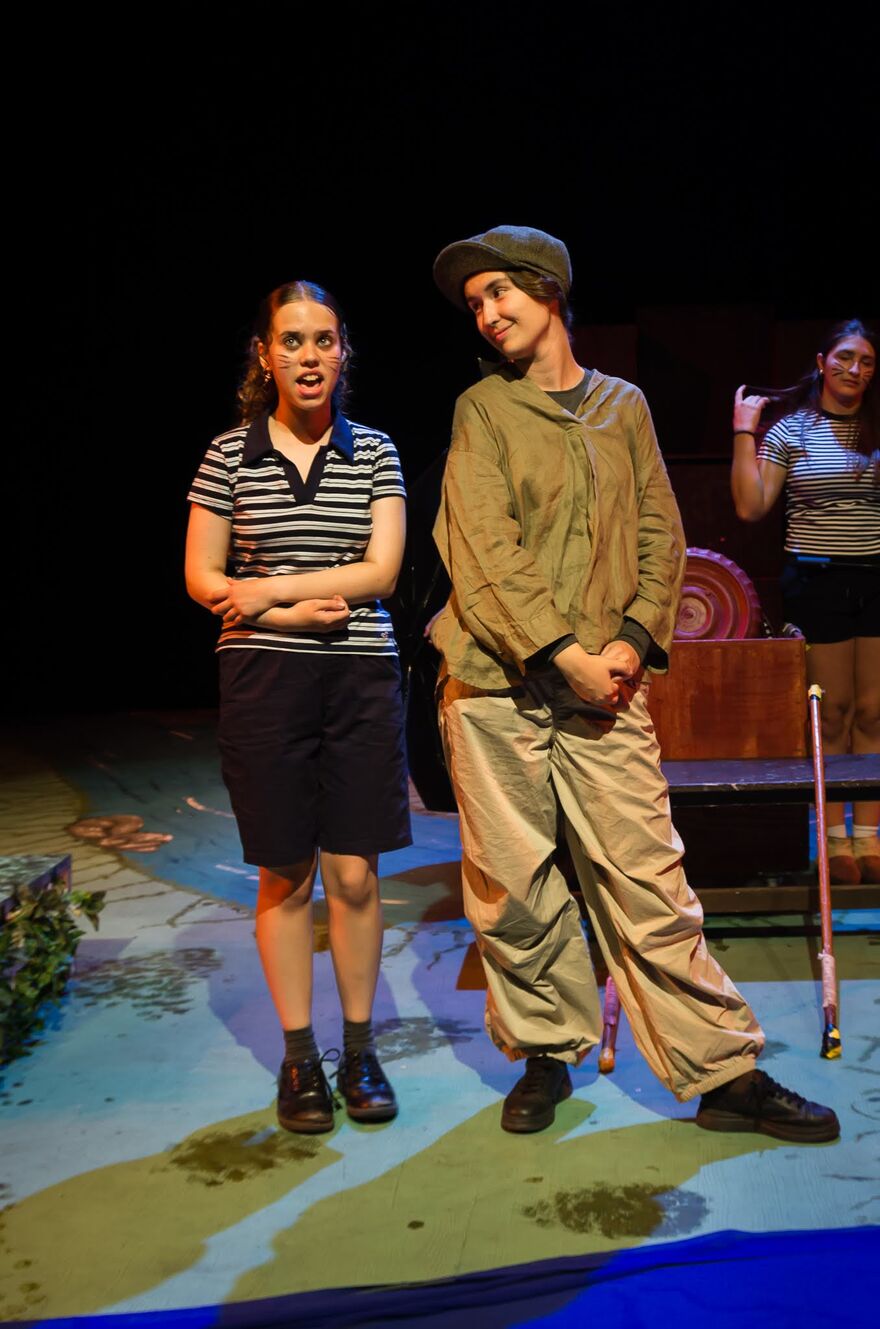two young people stand next to each other on stage. One is wearing stripes and the other is dressed in racing gear.