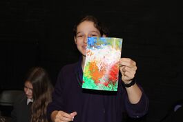 A young white women with long brown hair holds an A5 sheet of paper that has been coloured using red, orange, green, blue and yellow splatters of paint.