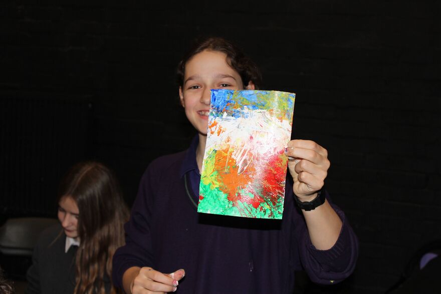 A young white women with long brown hair holds an A5 sheet of paper that has been coloured using red, orange, green, blue and yellow splatters of paint.