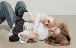 A white woman with blonde hair is lying on her back, cuddling a ventriloquist doll. She’s wearing a white shirt and jeans, and the doll is also blonde, with lined eyes and very red lips. It’s wearing a cream turtleneck jumper and chequered black and white trousers.