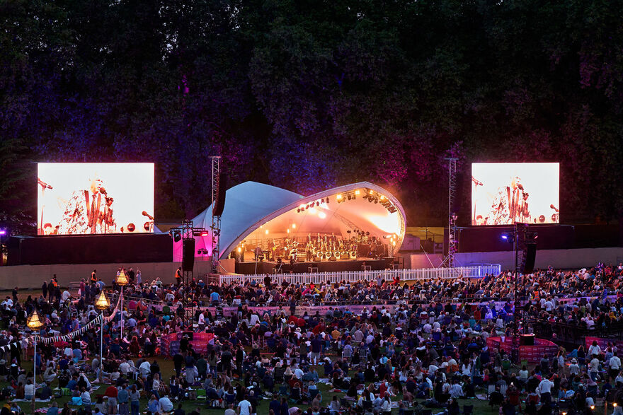 Round stage with orchestra and singers performing in Battersea Park, large crowd sits in front watching.
