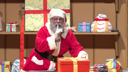 An actor dressed as Santa Claus has one finger to his lips, looking off to the side as his other hand holds a large present wrapped in a yellow bow. 