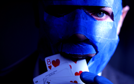 A white man with piercing blue eyes is mostly in shadow, with only the right edge of his face in the light. He has blue tape all over his face, covering everything but his eyes, nostrils and mouth. He is holding some playing cards to his mouth as if he is about to eat them, his hand is in a blue latex medical glove. 