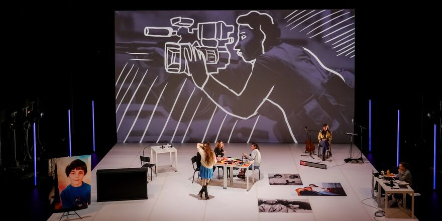 A large theatre studio space with a background photo image of a person with a gun, with an outline drawing of a person with a camera superimposed on the photo. The stage is populated by 5 performers of mixed gender. There are tables, chairs, microphones, a guitar, and large photo portraits of people spread around the stage.
