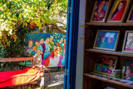 Photograph is taken from inside an artists studio looking out the door to the garden. The garden has a bench in the foreground and Lucinda Denning's brightly coloured canvas painting of a group of people in the background.